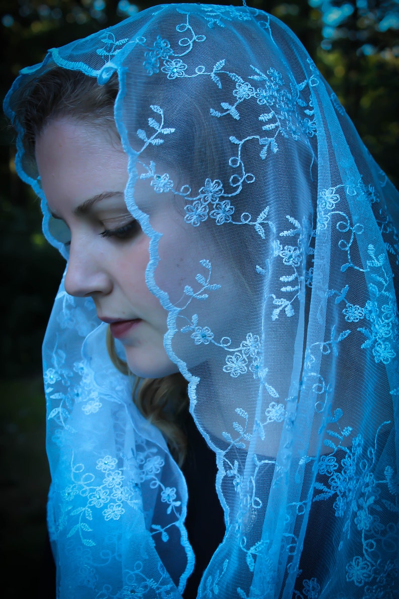 May include: A woman wearing a light blue lace veil with white floral embroidery. The veil covers her head and shoulders, and she is looking down.
