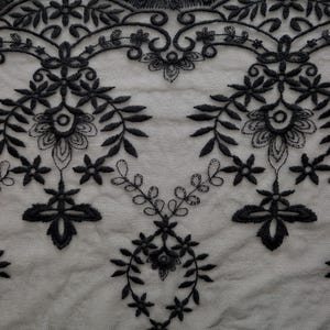 Puede incluir: Una tela de encaje negro con un intrincado patr&oacute;n floral bordado y un borde festoneado. El dise&ntilde;o consta de motivos bot&aacute;nicos sim&eacute;tricos, que incluyen hojas, peque&ntilde;as flores y enredaderas, cosidos sobre una base de malla transparente.