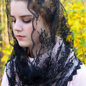 May include: A black lace veil with a floral pattern, draped over the head and shoulders. The veil is worn over a light pink sweater. The background features blurred yellow and green foliage.