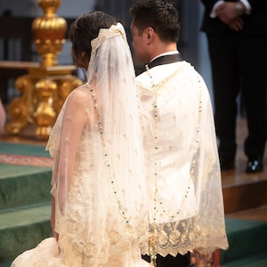 May include: A bride and groom at a wedding ceremony. The bride wears a white gown with lace detailing and a veil. The groom wears a white jacket with gold trim. Both are adorned with beaded necklaces.