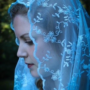 May include: A woman wearing a light blue lace veil with floral embroidery. The veil is sheer and delicate, and the floral pattern is intricate.