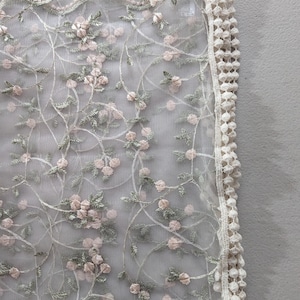 May include: Delicate floral lace fabric featuring a repeating pattern of pink blossoms and green leaves on a sheer white background. The edge is trimmed with a scalloped border and a cream-colored, textured trim. Ideal for bridal or craft projects.