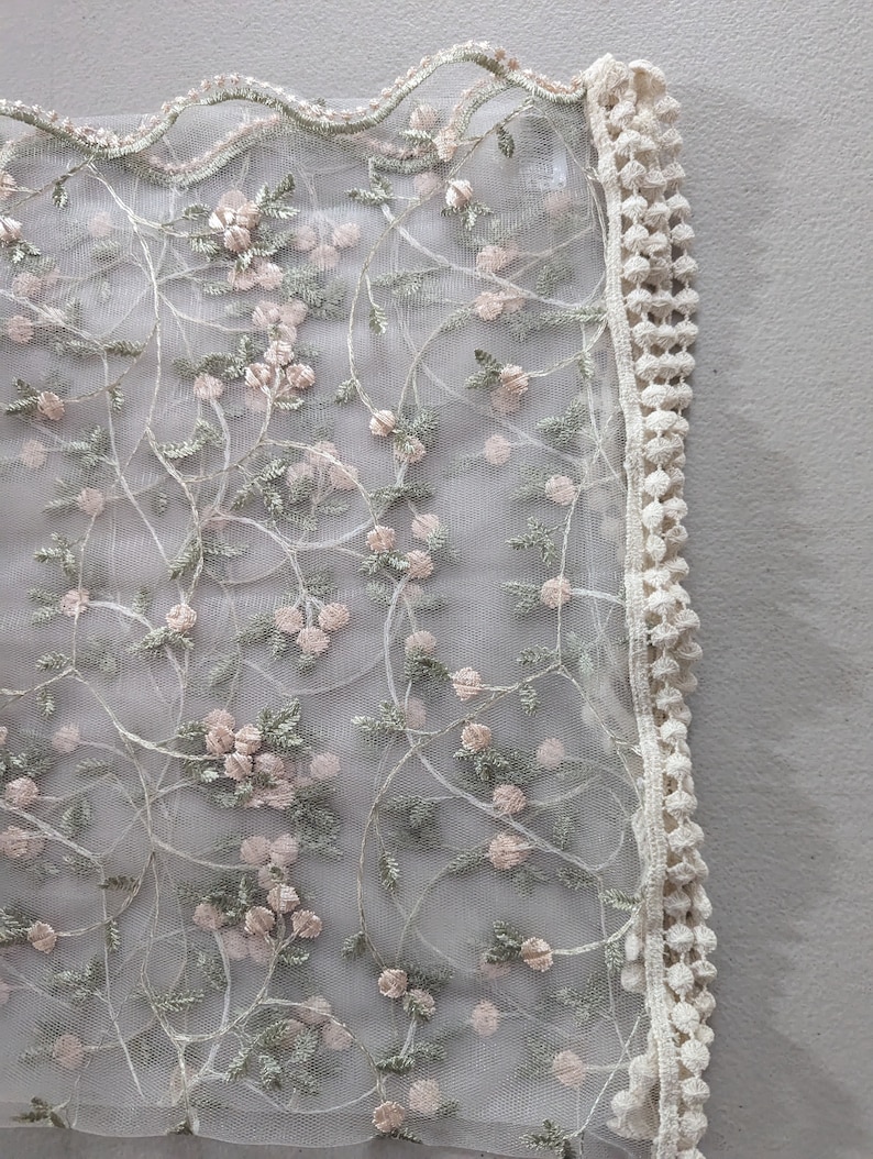 May include: Delicate floral lace fabric featuring a repeating pattern of pink blossoms and green leaves on a sheer white background. The edge is trimmed with a scalloped border and a cream-colored, textured trim. Ideal for bridal or craft projects.