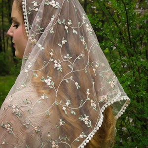 May include: A sheer, ivory-colored veil with delicate floral embroidery in pink and green. The veil features a scalloped edge with small, white pom-poms. The design is reminiscent of vintage bridal wear.