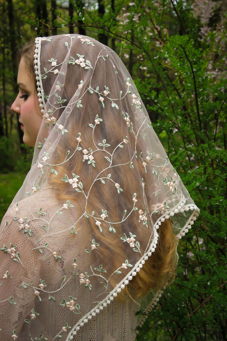 May include: A sheer, ivory-colored veil with delicate floral embroidery in pink and green. The veil features a scalloped edge with small, white pom-poms. The design is reminiscent of vintage bridal wear.