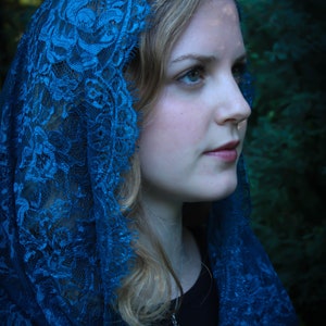 May include: A woman wearing a blue lace shawl with a silver pendant necklace. The shawl is draped over her head and shoulders, and she is looking off to the side.
