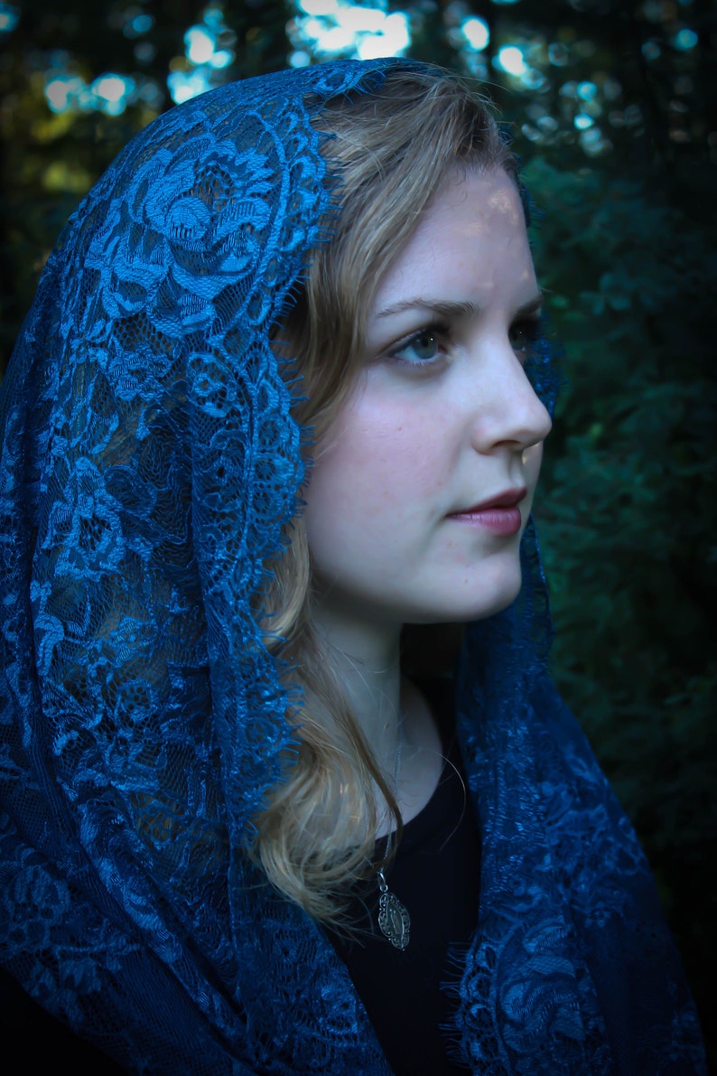 May include: A woman wearing a blue lace shawl with a silver pendant necklace. The shawl is draped over her head and shoulders, and she is looking off to the side.
