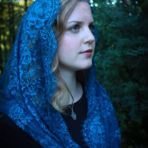 May include: A woman wearing a blue lace shawl over a black top. The shawl is draped over her head and shoulders, and she is looking off to the side.