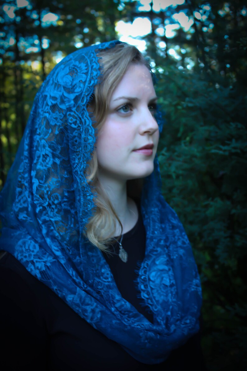 May include: A woman wearing a blue lace shawl over a black top. The shawl is draped over her head and shoulders, and she is looking off to the side.