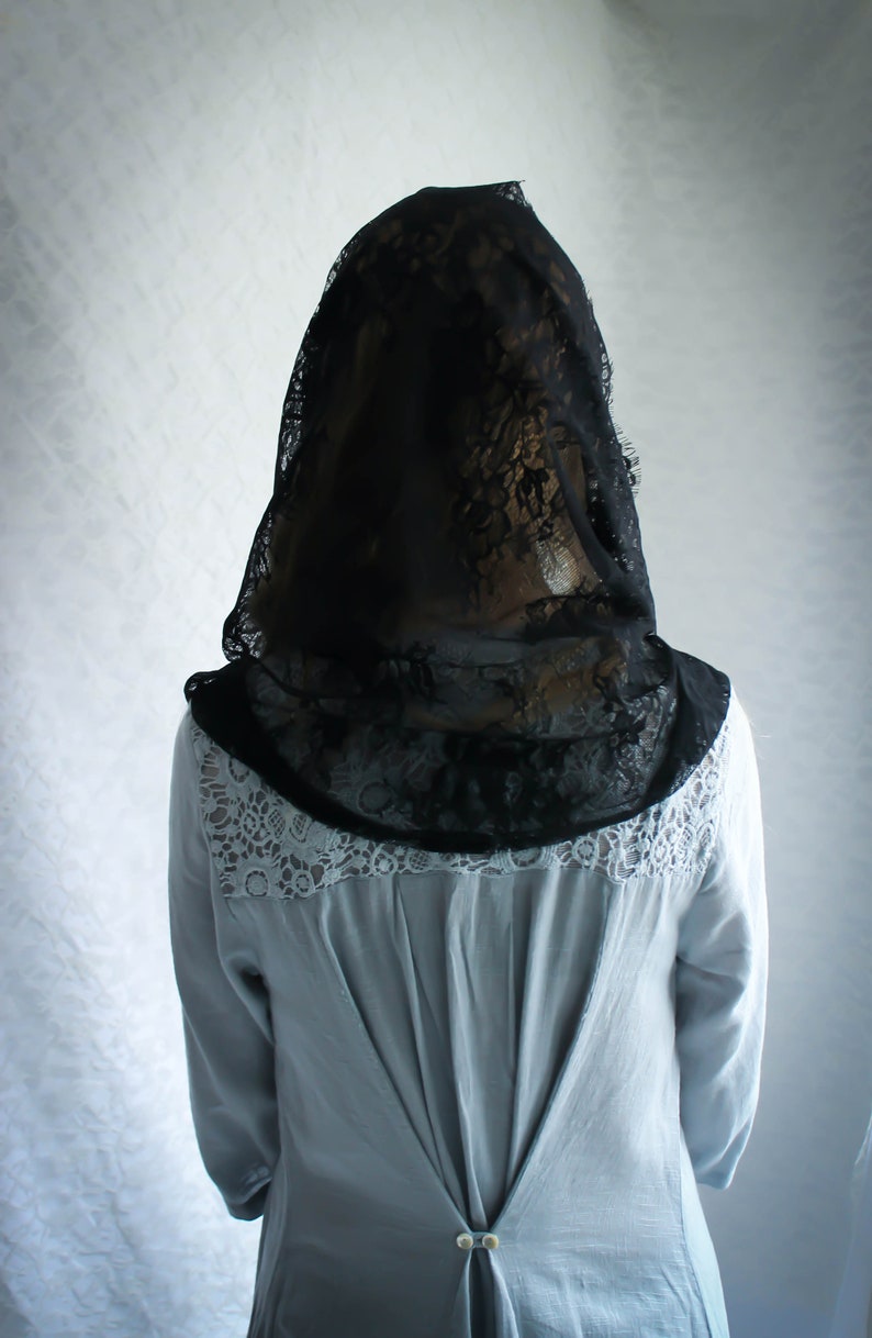 May include: A woman wearing a black lace veil over a light blue blouse with lace trim. The blouse has a button closure in the back.