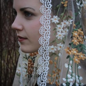 May include: A white lace veil with floral embroidery in shades of green, white, and yellow. The veil is sheer and delicate, with a scalloped edge.
