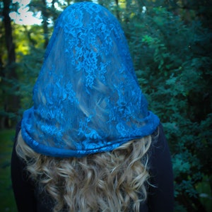 May include: A woman with long blonde hair is wearing a blue lace veil over her head. The veil is sheer and has a delicate floral pattern. The woman is standing in a forest setting.