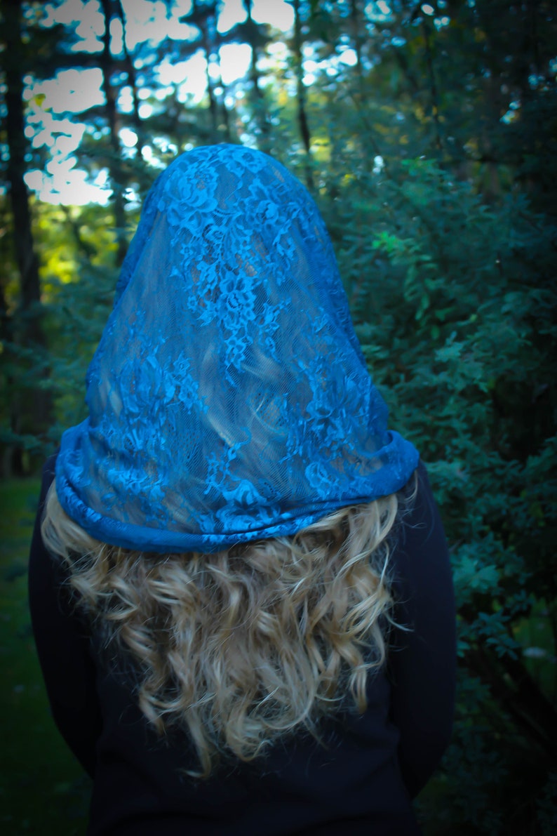 May include: A woman with long blonde hair is wearing a blue lace veil over her head. The veil is sheer and has a delicate floral pattern. The woman is standing in a forest setting.