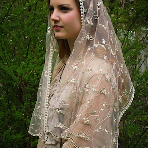 May include: A sheer, beige veil with delicate floral embroidery and a scalloped edge. The veil is adorned with small, white pearl-like beads along the border. The design features embroidered flowers in shades of pink and green.