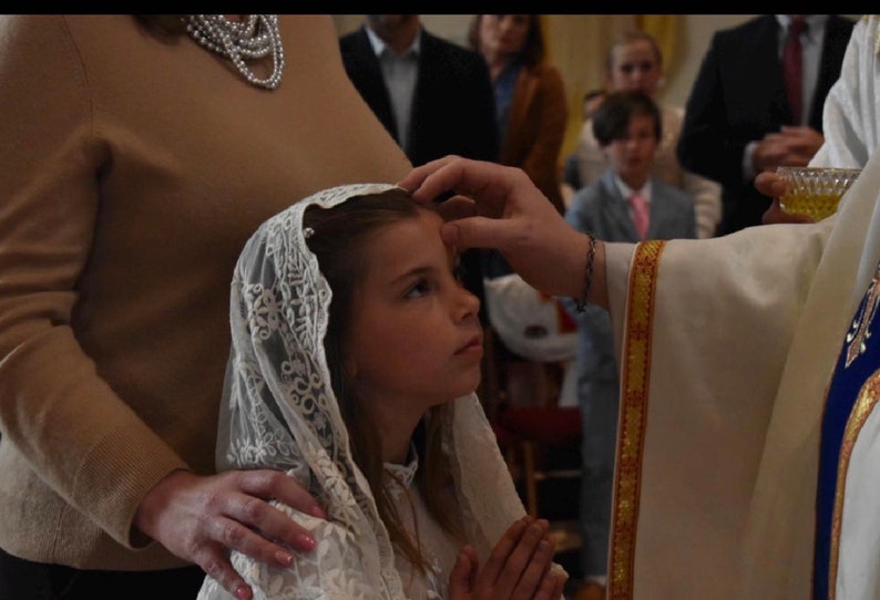 Puede incluir: Un velo de encaje blanco con intrincados bordados florales que lleva un ni&ntilde;o durante una ceremonia religiosa. La delicada tela cae sobre la cabeza y los hombros, presentando un borde festoneado y una base de malla fina con bordados detallados.