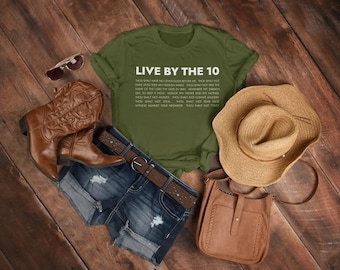 Könnte beinhalten: Ein grünes T-Shirt mit weißem Text, der "Live by the 10" lautet und die Zehn Gebote auflistet. Das Shirt liegt auf einer Holzoberfläche mit einem Paar braunen Cowboystiefeln, einem Jeansrock, einem Strohhut und einer braunen Ledertasche.