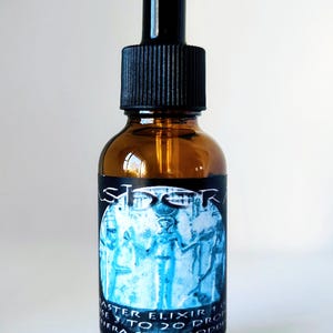May include: An amber glass dropper bottle with a black cap. The label features a blue-toned illustration of an Egyptian-style figure and the text: ASTER, MASTER ELIXIR, USE 3 TO 20 DROPS, HERA STAR GODDESS. The bottle has a cylindrical body and a dropper top.