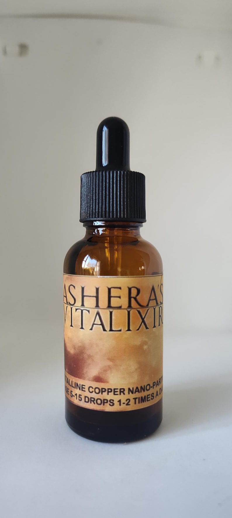 May include: An amber glass dropper bottle with a black cap. The label features the text ASHERA'S VITALIXIR in black lettering, with additional text reading CRYSTALLINE COPPER NANO-PARTICLES and USE 5-15 DROPS 1-2 TIMES A DAY on a tan background.