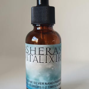 May include: An amber glass dropper bottle with a black cap. The label features the text SHERA'S VITALIXIR, followed by the text ALLINE SILVER NANO-PARTICLES and 15 DROPS 1-2 TIMES A DAY, set against a blue and white cloudy background design.
