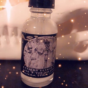 May include: A clear glass bottle with a black dropper top and a label that reads "Ethers Elixir Use 3 to 20 Drops Ashera Star Coven". The bottle is sitting on a white surface with a blurry background of golden lights.