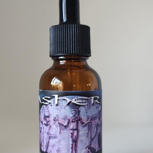 May include: An amber glass dropper bottle with a black cap. The label features a purple-toned illustration of an ancient figure and the text: ASHERA, REAL EVOLUTION ELIXIR, USE 8-15 DROPS, ASHERA STAR GODDESS. The bottle is cylindrical with a tapered neck.