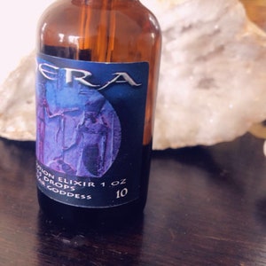 May include: A brown glass bottle with a silver dropper top and a blue label. The label has a purple image of two figures and the text "UTION ELIXIR 1 oz 10 DROPS TAR GODDESS".