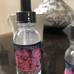 May include: A clear glass bottle with a black dropper top and a label that says "Isis Potion Elixir 3 to 20 Drops A Star Concoction". The label features a pink image of a woman in a robe with a crown on her head.