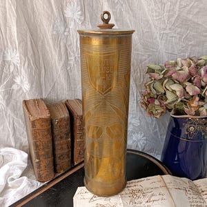 May include: A brass cylinder with a lid, engraved with the words "Verdun Meuse 15-16" and a shield design. The cylinder is decorated with a repeating pattern of birds and flowers.