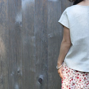 May include: A light beige linen top with short sleeves and a scoop neckline. The top is paired with a floral print skirt. The background is a weathered wooden fence.