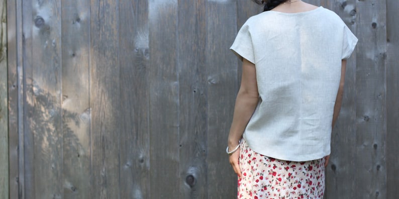 May include: A light beige linen top with short sleeves and a scoop neckline. The top is paired with a floral print skirt. The background is a weathered wooden fence.
