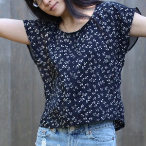 May include: A dark blue top with a white floral pattern. The top has short, flowing sleeves and is paired with denim shorts. The background is a wooden fence.