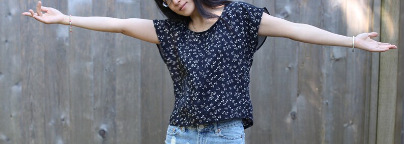 May include: A dark blue top with a white floral pattern. The top has short, flowing sleeves and is paired with denim shorts. The background is a wooden fence.