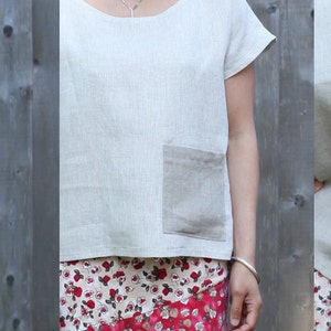May include: A light beige linen top with short sleeves and a round neckline. The top features a patch pocket on the front and a contrasting panel across the back. The fabric has a slightly textured appearance. The top is paired with a floral print skirt.