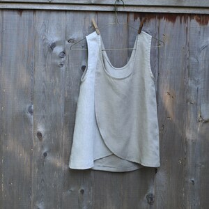 May include: A light gray linen sleeveless top with a wrap-style design. The top is hanging on a wooden hanger against a gray wooden background.