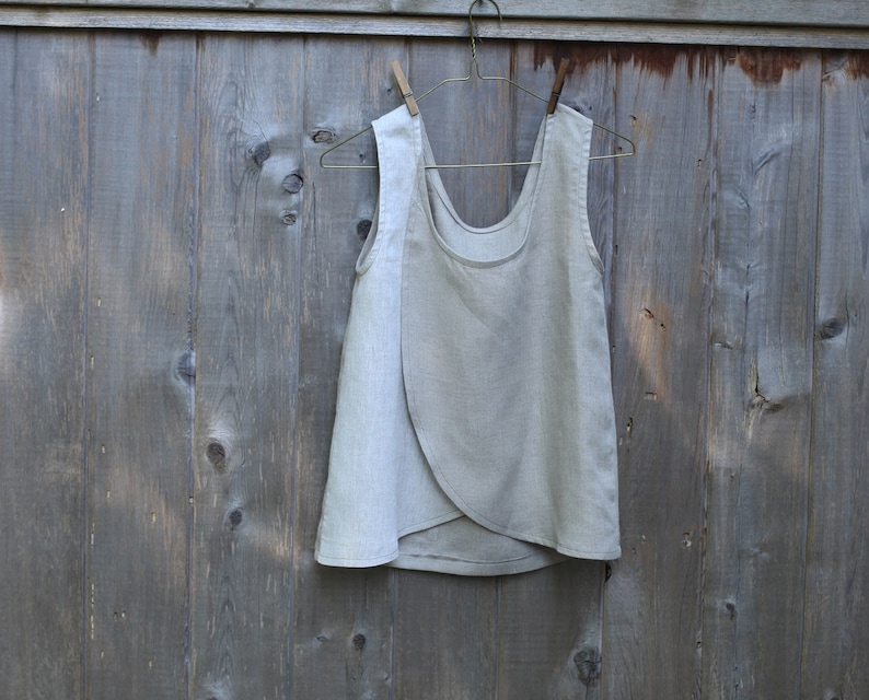 May include: A light gray linen sleeveless top with a wrap-style design. The top is hanging on a wooden hanger against a gray wooden background.