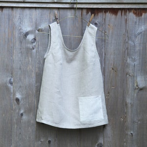 May include: A light beige linen apron with a single pocket on the front. The apron is hanging on a wooden hanger with two clothespins.