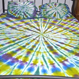 May include: A tie-dye bedspread with a circular pattern in shades of green, blue, purple, and yellow. The bedspread is on a bed with two matching tie-dye pillowcases.