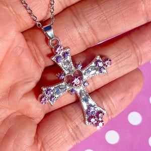 Könnte beinhalten: Halskette mit silberfarbenem Kreuzanhänger und feiner Kette. Das Kreuz ist mit klaren und lila Edelsteinen besetzt, darunter ein herzförmiger Mittelstein. Der Anhänger wird in einer Hand vor einem rosa Hintergrund mit weißen Punkten gehalten.