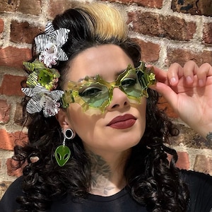 May include: Translucent lime green sunglasses with a jagged, star-like frame edge. The eyewear is paired with a dangling green alien head earring and a hair accessory featuring white and black zebra-striped flowers with green and white orchid accents.