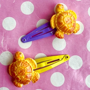 Op de afbeelding: Twee haarclips met schildpadontwerpen. Eén clip is paars, de andere is geel. De schildpadden zijn goudbruin. De achtergrond is roze met witte stippen.