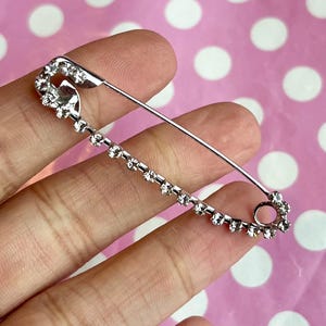 Może przedstawiać: Srebrna broszka w kształcie agrafki z dekoracyjnym wzorem. Szpilka posiada rząd małych, przezroczystych kryształków na jednej stronie i dekoracyjny wierzch z większą ilością kryształków. Broszka trzymana jest w dłoni.