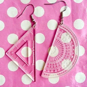 Puede incluir: Un par de pendientes rosas de acrílico. Un pendiente es una regla triangular, el otro es un transportador. El texto "Tomorrow is better" está impreso verticalmente en el pendiente de la regla. Los pendientes cuelgan de ganchos plateados.
