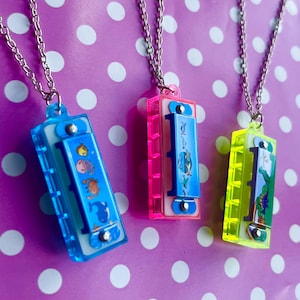 May include: Three harmonica pendant necklaces with silver chains. The harmonicas are in blue, pink, and yellow, each with a different design. The background is a purple surface with white polka dots.