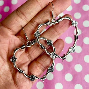 Puede incluir: Pendientes de aro de plata con diseño de alambre de púas y detalles de rosas. Los pendientes son grandes y tienen forma de corazón.