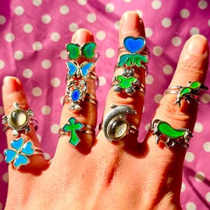 May include: A hand displays a collection of silver rings with various designs. The rings feature a four-leaf clover, a cross, butterflies, a dolphin, and other shapes, with blue and green enamel accents. The background is a pink polka dot pattern.