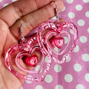 May include: Pink heart-shaped earrings with a layered design. The earrings are made of clear plastic with a pink tint. The hearts are layered on top of each other, creating a three-dimensional effect.
