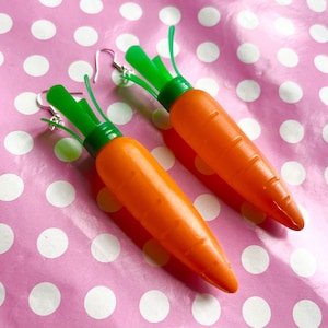 May include: A pair of novelty carrot earrings. Each earring features a bright orange carrot with green tops and silver-coloured ear wires. The earrings are set against a pink background with white polka dots.
