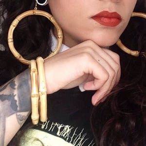 Op de afbeelding: Een vrouw met donkerbruin haar en een enkele blonde streep in haar haar draagt grote, ronde, houten oorbellen. Ze draagt ook een zwart shirt en heeft een tatoeage op haar arm. Ze heeft een rode roos in haar haar.