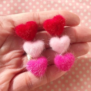 Könnte beinhalten: Ein Paar flauschige Herzohrringe in Pink, Rot und Hellrosa. Die Ohrringe sind übereinander gelegt, wobei das größte Herz unten und das kleinste Herz oben ist.
