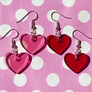 May include: Three pairs of heart-shaped earrings. One pair is red, one pair is pink, and one pair is a gradient of pink and red.